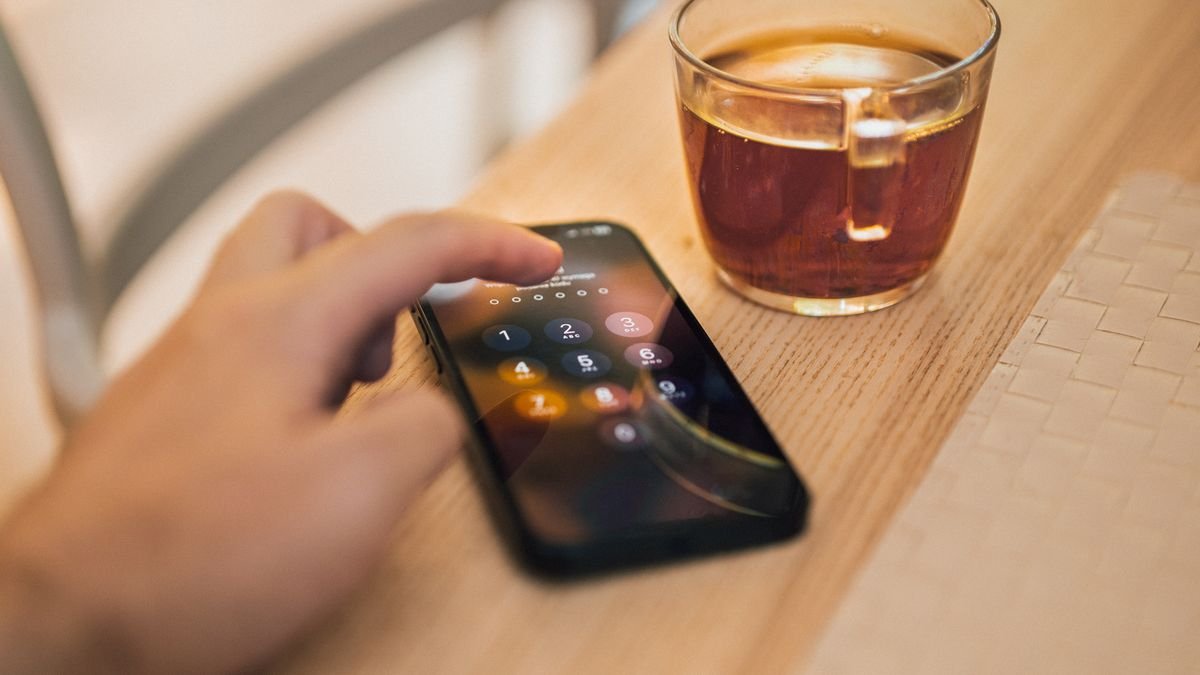 Tangan membuka kunci layar ponsel di samping secangkir teh di atas meja kayu